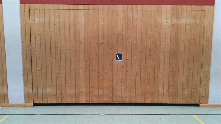 Einzelnes geschlossenes Geräteraumtor aus senkrechten Holzlamellen mit eingelassenem Muschelgriff in einer Sporthallenwand