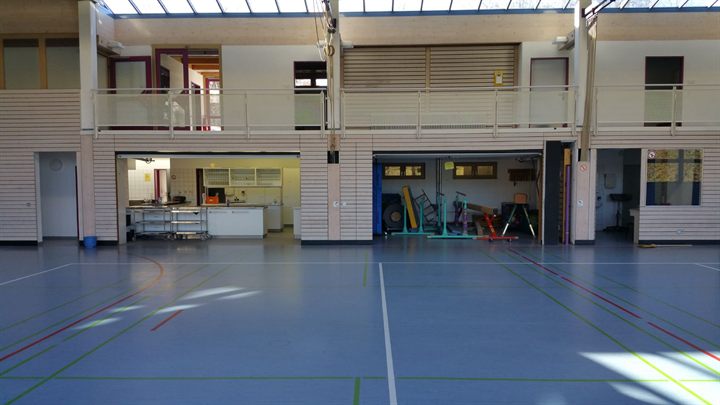Sporthalle mit zwei geöffneten Geräteraumtoren; im Geräteraum stehen Barren, Sprungkasten und weitere Turngeräte