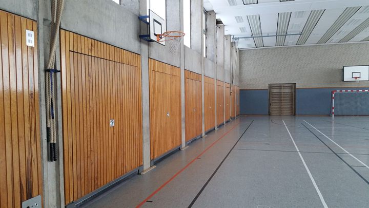 Sporthalle mit einer Reihe geschlossener hölzerner Geräteraumtore zwischen Betonstützen entlang der Längsseite